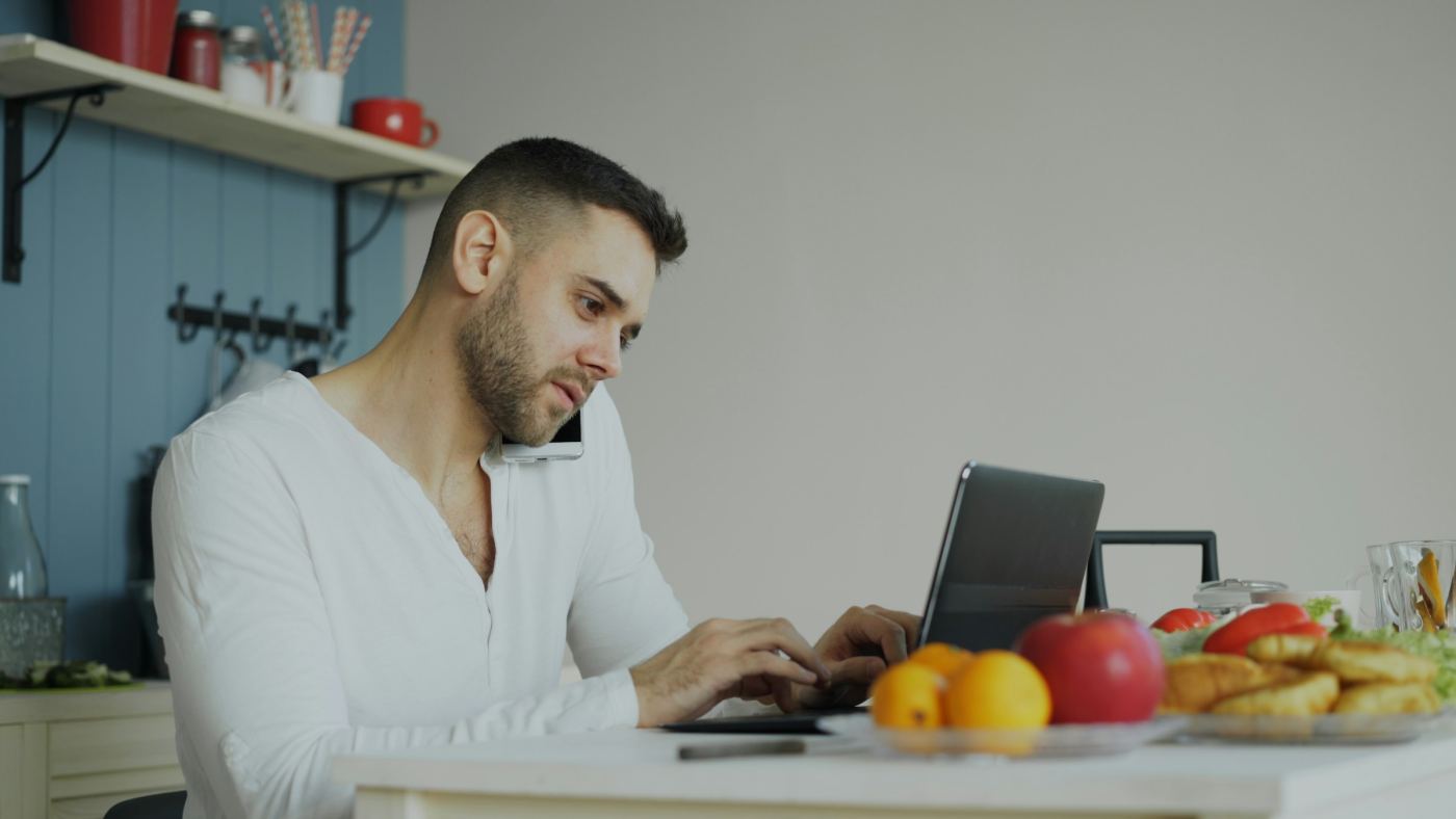 Een man aan de keukentafel met een laptop en een telefoon.